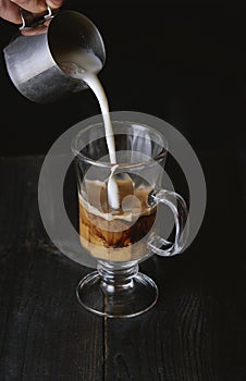 Hand pouring milk in cold brew coffee on black wooden table.