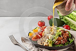 A hand pouring salad dressing into cobb salad with chicken, avocado, bacon, blue cheese, tomato and eggs. Keto diet