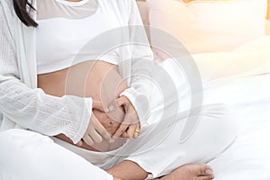 Hand of pregnant woman in white dress make heart with hand on her belly in bedroom. Make heart-shaped symbol on the belly of