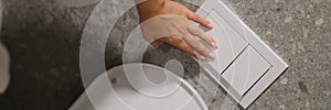 Hand pressing button to drain water in toilet bowl in restroom closeup