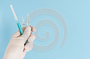 Hand in protective glove holding ampules with vaccine and syringe on blue background