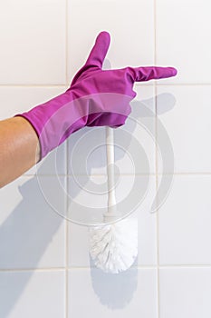 Hand in a purple glove holds a white toilet brush against a background of white tiles