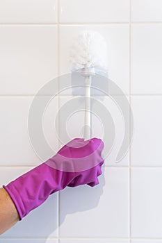 Hand in a purple glove holds a white toilet brush against a background of white tiles