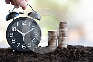 Hand put black clock on soil with stack of coin. Time to save money concept. Coins with a backdrop nature blurred