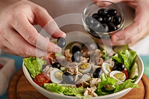 Hand put olive into the bowl with fresh vegetables and canned tuna.