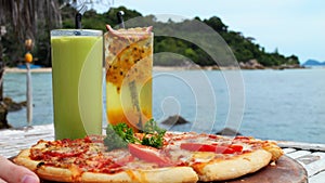 A hand puts a wooden tray with pizza on the table. Green smoothie with ice cubes and a drink with a slice of passion