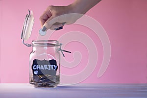 Hand putting coin in a glass jar with coins with a chalk Charity tag on a pink background. Donation and charity concept