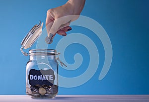 Hand putting a coin in a glass jar with coins with a chalk donate tag on a blue background. Donation and charity concept