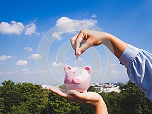 Hand putting coin into pink piggy bank with blue sky background, saving money for future investment plan and retirement fund