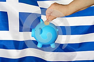 Hand putting coin to piggy bank on Greece flag background