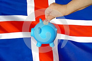 Hand putting coin to piggy bank on Iceland flag background