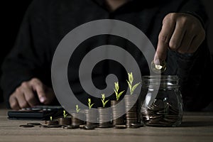 Hand putting coin to saving jar and coins stacking with plant growth on table. Business investment growth and profit increase