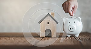 Hand putting coin on White piggy bank with wooden house on wood table and blurred copy space background. Concept for financial hom