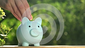 Hand putting coins into piggy bank on green nature background in rays of sun