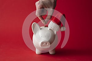 Hand putting money into a piggy bank wearing a santa hat. Christmas savings background