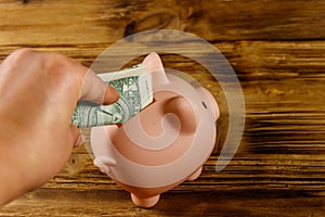 Hand putting one dollar bill into pink piggy bank. Saving money concept