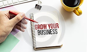 Hand with red pen. Cofee cup. Stick. Keyboard and white background. GROW YOUR BUSINESS sign in the notepad