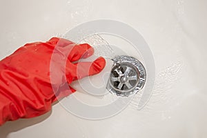 A hand in a red rubber glove with a melamine sponge rubs the white bathroom near the drain hole where the water flows