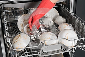 Hand in a red rubber glove puts a glass and dirty dishes in the dishwasher