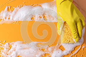 A hand in a rubber glove holds a cleaning sponge with foam on a bright yellow background. household chores and cleaning concept