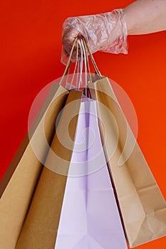 A hand in rubber or plastic glove holds paper eco-craft bags on red background. Gifts during coronavirus. Virus protection, social