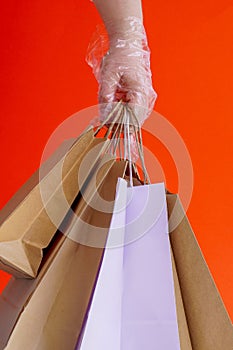 A hand in rubber or plastic glove holds paper eco-craft bags on red background. Gifts during coronavirus. Virus protection, social