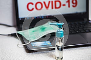 Hand sanitizer bottle and surgical mask on a laptop keyboard that reads covid-19 on the screen. Illustration to protect office