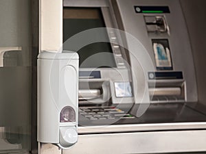 Hand sanitizer dispenser mounted on an ATM distributing cash money in fron of a bank, full of disinfecting hydroalcoholic gel