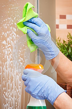 Hand of senior woman cleaning glass shower in bathroom using microfiber cloth and detergent