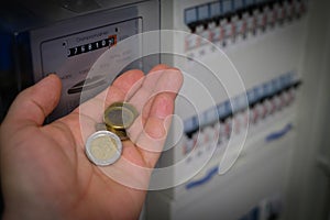 In a hand are several euro coins in front of a three-phase meter