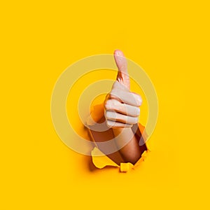 Hand showing a thumb up sign through a ripped hole in yellow paper background.