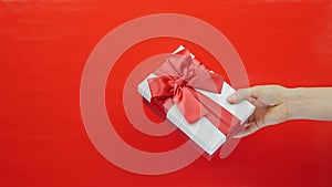 Hand is showing white box with red ribbon bow on red background. Female is holding valentines day gift in hand. St. Valentine`s Da