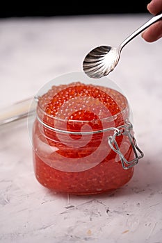 Hand with a spoon with red caviar over a glass jar filled with red caviar of salmon fish on a light background. Seafood, caviar