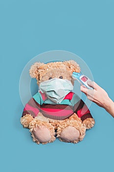 Hand with thermometer and teddy bear in mask on blue