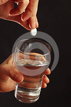 Hand throwing an effervescent pill in a glass of water on dark background.