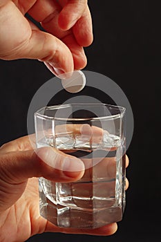 Hand throwing an effervescent pill from headache in glass of water on a dark background.