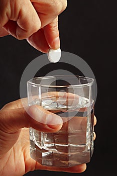 Hand throwing an effervescent tablet from headache in a glass of water on dark background.