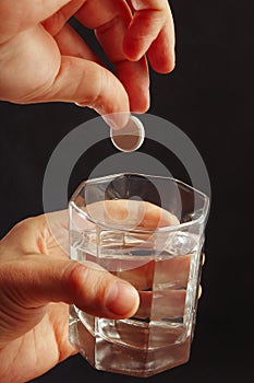 Hand throwing a soluble pill from headache in a glass of water on dark background.