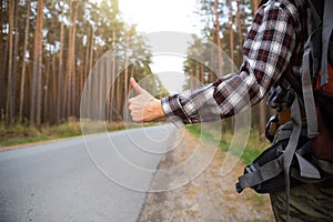 Hand with thumbs up on the background of highway - hitchhiking, voting. Tourist in a check shirt with backpack near the forest