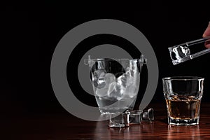 Hand with tongs holding ice-cube above a glass with whiskey, ice bucket behind the alcoholic drink on wooden table in restaurant.