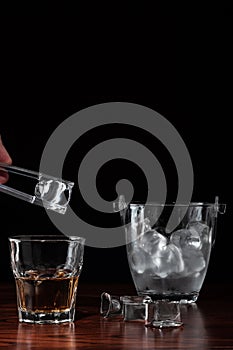 Hand with tongs holding ice-cube above a glass with whiskey, ice bucket behind the alcoholic drink on wooden table in restaurant.
