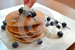 hand topping pancake stack with blueberries, yogurt on side