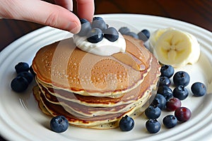 hand topping pancake stack with blueberries, yogurt on side