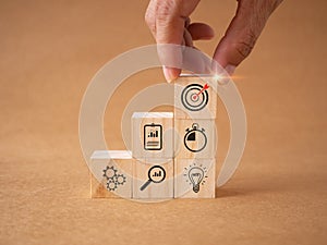 Hand touch wooden cubes stacking with icons goal, business strategy on brown workplace background.