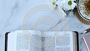 Hand turning pages on open holy bible book with cup of coffee and flowers on white marble background