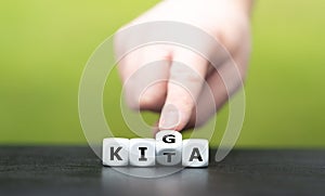 Hand turns dice and changes the German abbreviation KITA daycare to KIGA Kindergarten.