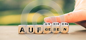 Hand turns dice and changes the German expression `aufgeben` `give up` to `aufstehen` `get up`.