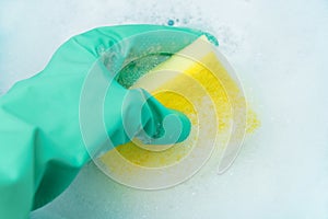 A hand in a turquoise glove holds a sponge for washing dishes in a copious foam.