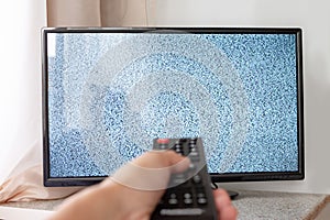 Hand with TV remote control in front of the screen with white noise on it - tuning the television channels and connecting problems