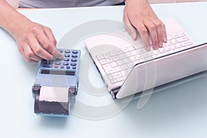 Hand typing on a laptop and a cash register on a blue background. Sales person entering amount on cash register